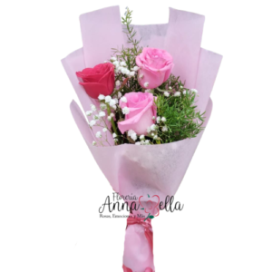 "Ramo de mano compacto con tres rosas naturales de diferentes colores delicados, acompañadas de follaje verde y envueltas en papel decorativo con un lazo."
