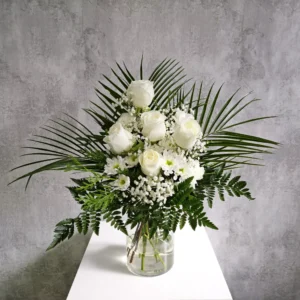 Arreglo floral en abanico dentro de un florero cilíndrico de vidrio transparente con agua. El ramo tiene cinco rosas blancas en el centro, margaritas blancas y gipsofila, enmarcado por hojas verdes de palma extendidas y helechos en la base, sobre una superficie blanca y fondo gris texturizado.
