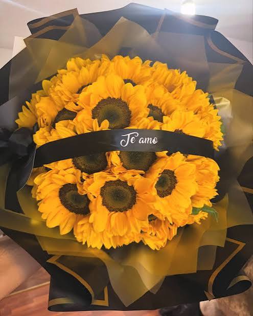 Un ramo grande y denso compuesto por veinte girasoles de color amarillo brillante con centros oscuros. El ramo incluye una cinta decorativa cruzada con una dedicatoria personalizada impresa sobre ella.
