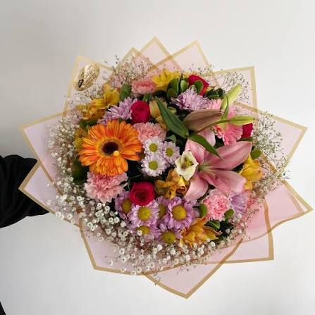 Vista superior de un ramo de flores grande y redondo. Contiene una gerbera naranja, lirios rosa, rosas rojas, claveles rosa, crisantemos morados y nubes de gypsophila blanca. El ramo está envuelto en papel coreano rosa con bordes dorados en forma de estrella.
