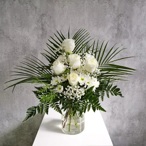 Arreglo floral en abanico dentro de un florero cilíndrico de vidrio transparente con agua. El ramo tiene cinco rosas blancas en el centro, margaritas blancas y gipsofila, enmarcado por hojas verdes de palma extendidas y helechos en la base, sobre una superficie blanca y fondo gris texturizado.