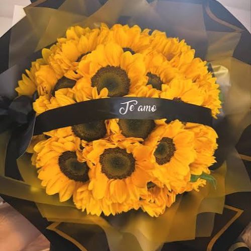 Un ramo grande y denso compuesto por veinte girasoles de color amarillo brillante con centros oscuros. El ramo incluye una cinta decorativa cruzada con una dedicatoria personalizada impresa sobre ella.